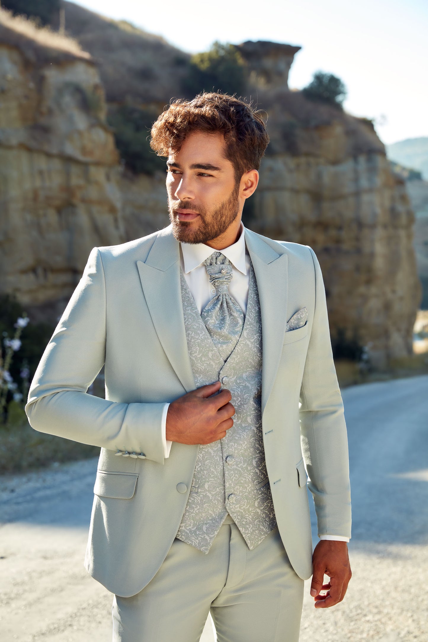 Homme qui porte un costume de marié de coloris vert eucalyptus. Le costume est un costume trois pièces avec veste et pantalon uni et un gilet et une cravate aux motifs floraux dans une teinte plus claire.