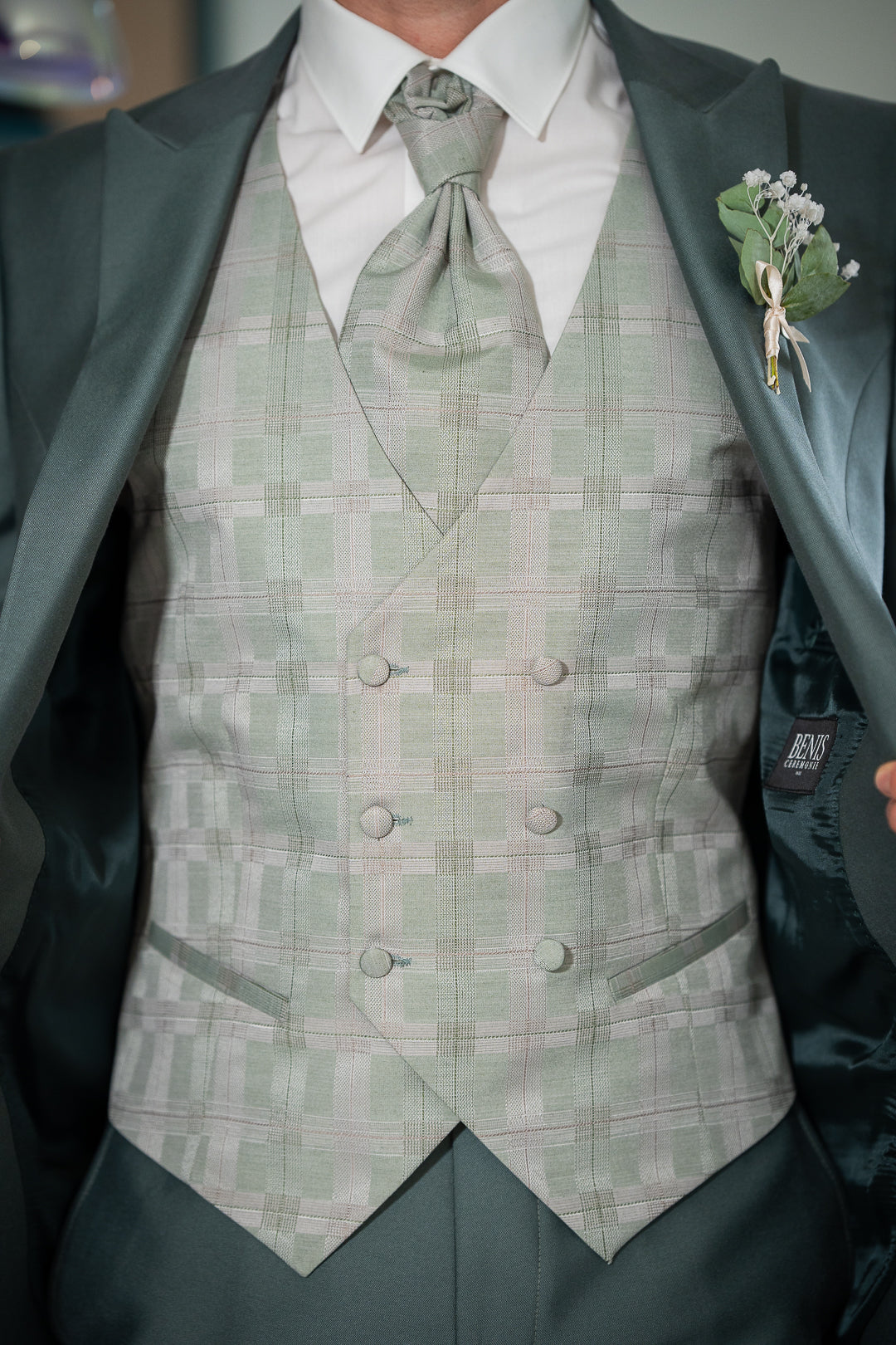 L'image montre un homme portant un costume élégant. La veste est vert foncé et le gilet est vert clair à carreaux. La cravate est assortie au gilet. Le costume est de haute qualité et bien taillé. La boutonnière est ornée d'une petite fleur blanche. L'ensemble est très chic et parfait pour un mariage