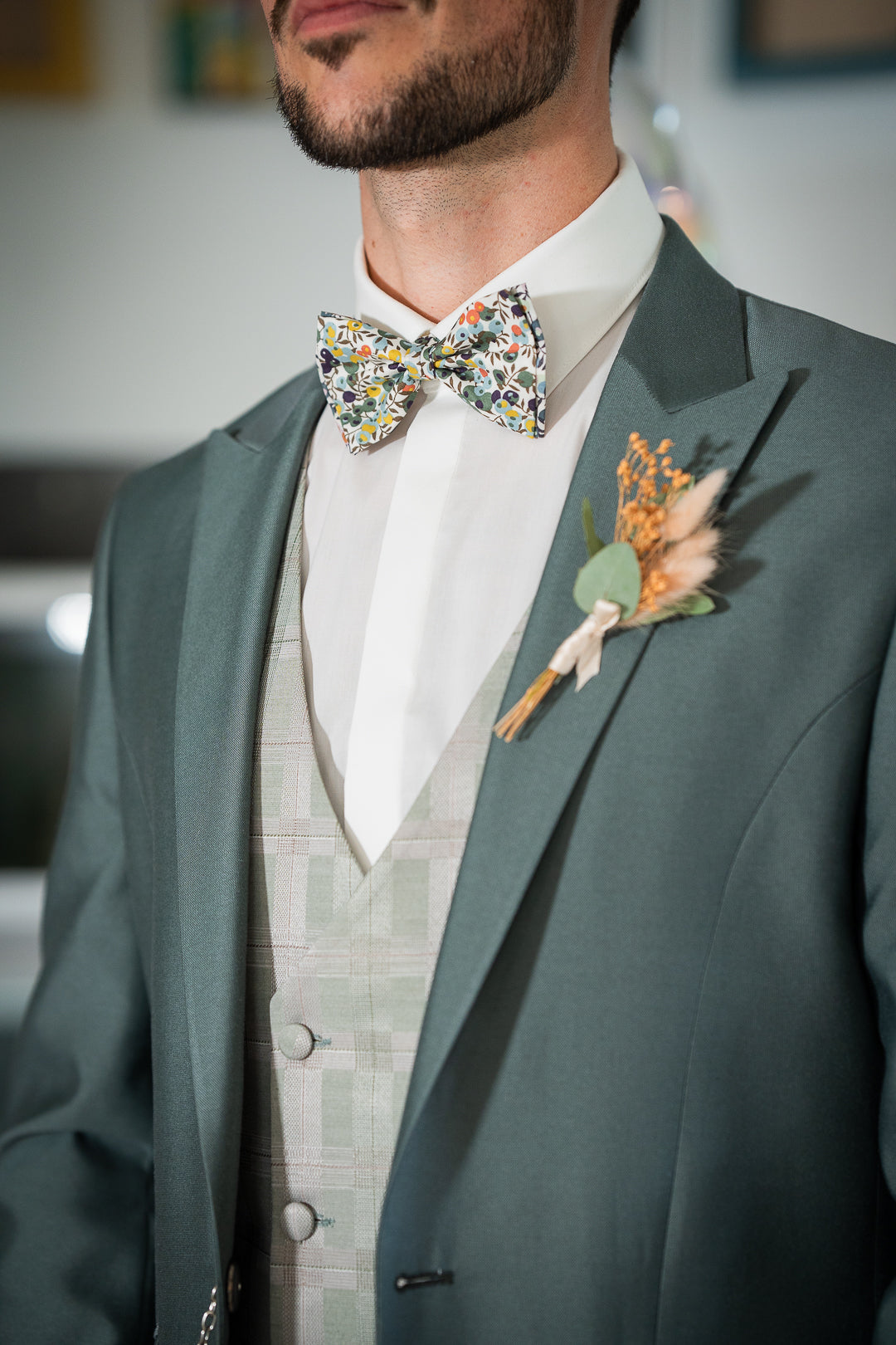 L'image montre un homme portant un costume vert élégant pour un mariage. Le costume est composé d'une veste vert sauge, d'un gilet à carreaux beige et blanc cassé, et d'une chemise blanche classique. Le détail le plus accrocheur est le nœud papillon à fleurs, qui apporte une touche de couleur et de fantaisie à l'ensemble. La petite boutonnière florale ajoute une touche finale raffinée. L'ensemble est à la fois moderne et chic, parfait pour une cérémonie formelle.
