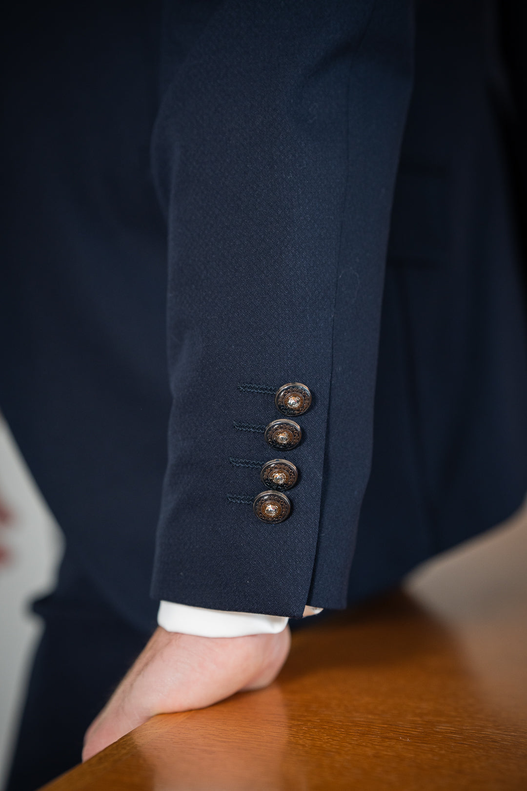 L'image montre un gros plan sur la manche d'une veste de costume bleu marine. La manche est ornée de quatre boutons décoratifs. Une main masculine repose sur une table en bois. Le tissu de la veste semble être de haute qualité, avec une texture fine et soignée. La couleur sombre et la qualité du tissu suggèrent un vêtement formel, peut-être porté pour un mariage