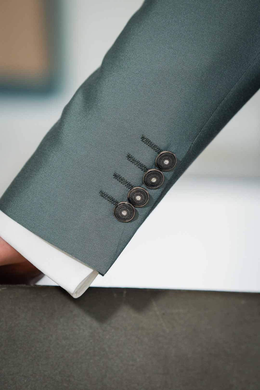 L'image montre un détail d'une manche de veste de costume verte. Le tissu est de couleur vert sauge, avec une texture fine et soignée. La manche est ornée de quatre boutons de manchette foncés, métalliques, ornés de petits détails brillants, probablement des cristaux ou des pierres. Les boutons sont attachés à la manche par des fils noirs fins, presque invisibles. On aperçoit également un petit morceau de la chemise blanche sous la manche du costume.