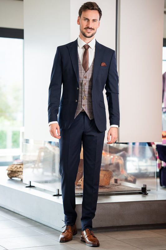 L'image montre un jeune homme élégant portant un costume bleu marine. Le costume est composé d'une veste et d'un pantalon assortis. Il porte également une veste de gilet à carreaux gris et brun clair par-dessus une chemise blanche. Sa cravate est brun foncé, assortie à la couleur de son gilet. Ses chaussures sont en cuir brun clair.
