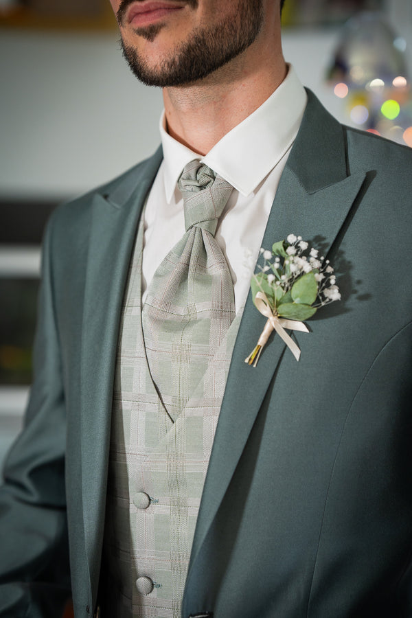 L'image montre le haut du corps d'un homme vêtu d'un costume vert-gris élégant. Le costume est composé d'une veste, d'un gilet et d'une chemise blanche. La cravate est assortie au gilet, présentant un motif à carreaux vert pâle et beige. Une petite boutonnière, composée de petites fleurs blanches et de feuilles vertes, est épinglée sur la veste, ajoutant une touche délicate à l'ensemble. Le style général est formel et soigné, suggérant peut-être une occasion spéciale comme un mariage.