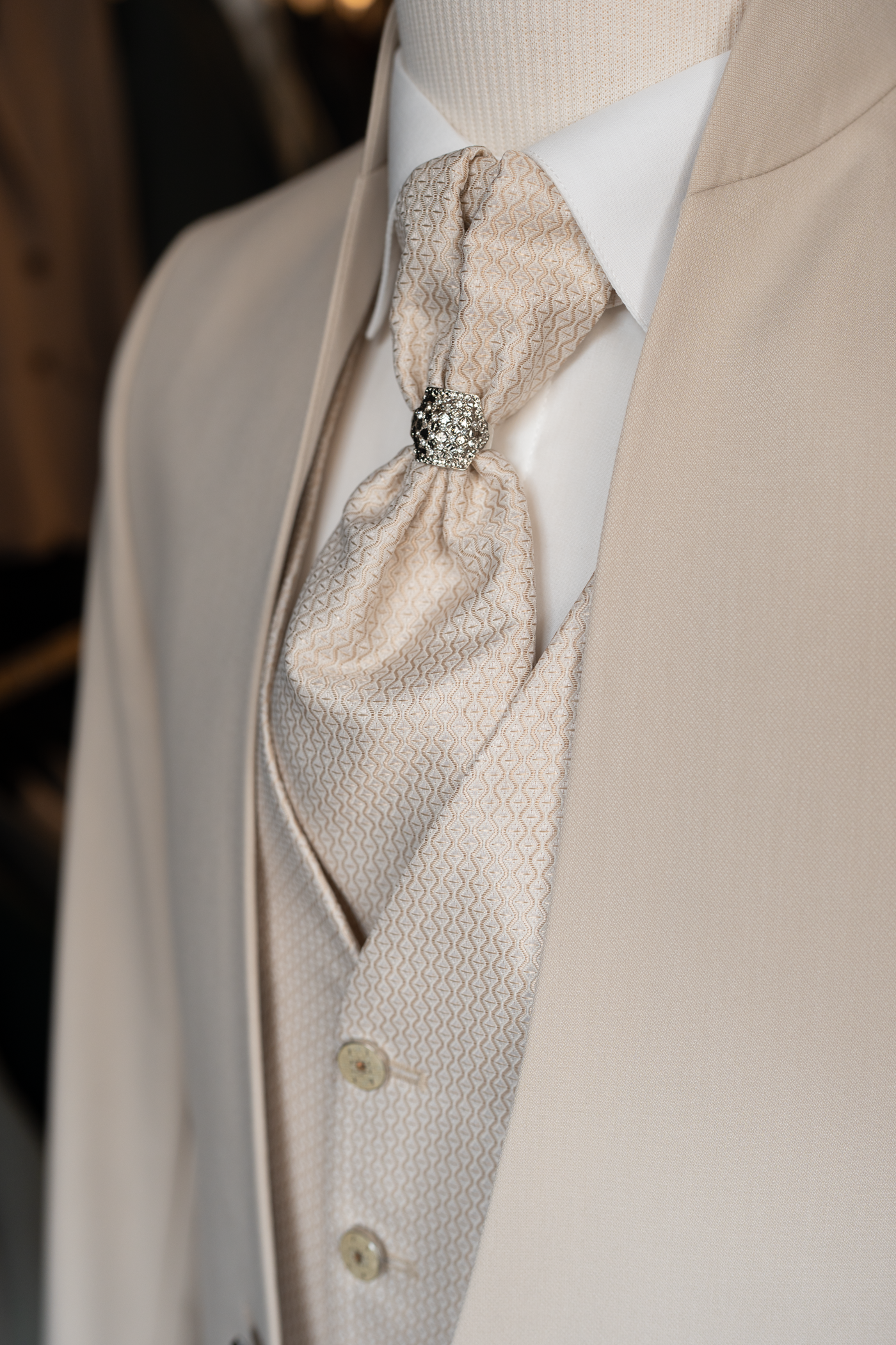 Vue rapprochée d'un ensemble de mariage haut de gamme pour homme en beige clair.
Le gilet et la cravate plastron assortie présentent un motif tissé géométrique sophistiqué.
Un bijou de cravate argenté serti de cristaux maintient le nœud de la cravate plastron.
La veste à col mao encadre la tenue, révélant une chemise blanche immaculée en dessous.
Les boutons du gilet sont assortis au tissu beige, complétant ce look de cérémonie raffiné.