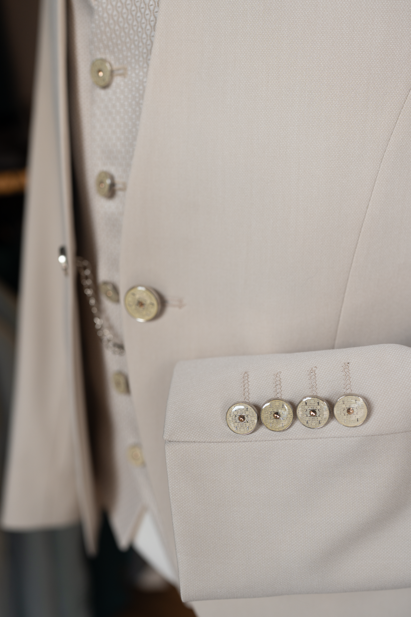 Gros plan détaillé sur le poignet d'une veste de costume de mariage pour homme, de couleur beige clair. Quatre boutons de manchette ronds et décoratifs, chacun orné d'un motif complexe et d'un petit élément doré ou ambré au centre, sont alignés sur la manche. Les boutonnières sont cousues avec soin en fil de couleur assortie. Le tissu texturé de la veste est clairement visible. L'arrière-plan, légèrement flou, laisse entrevoir d'autres parties du costume, y compris le gilet assorti et une petite chaîne déco