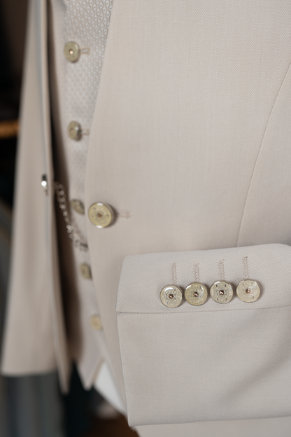 Gros plan détaillé sur le poignet d'une veste de costume de mariage pour homme, de couleur beige clair. Quatre boutons de manchette ronds et décoratifs, chacun orné d'un motif complexe et d'un petit élément doré ou ambré au centre, sont alignés sur la manche. Les boutonnières sont cousues avec soin en fil de couleur assortie. Le tissu texturé de la veste est clairement visible. L'arrière-plan, légèrement flou, laisse entrevoir d'autres parties du costume, y compris le gilet assorti et une petite chaîne déco