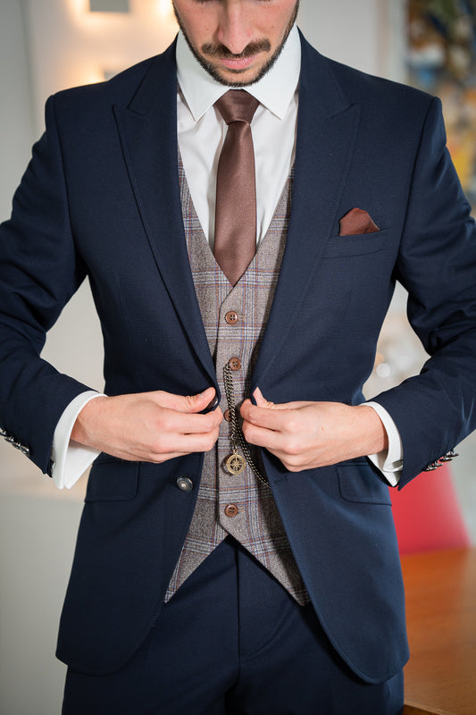 L'image montre un homme vêtu d'un costume bleu marine élégant. Le costume comporte un gilet à carreaux gris et brun clair assorti à une cravate brune. L'homme ajuste sa montre de poche, accrochée à sa veste par une fine chaîne.  Le détail et la composition de l'image suggèrent une attention particulière aux détails et un style rétro et vintage. L'ensemble est formel et classique.

