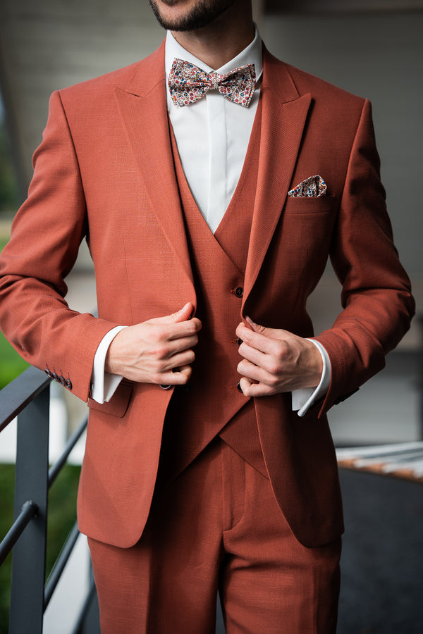 L'image montre un homme portant un costume trois pièces terracotta.
Le costume est élégant et bien taillé, avec une coupe ajustée. Le veston possède une poche poitrine dans laquelle se trouve une pochette de couleur claire à motifs floraux. L'homme porte également un nœud papillon assorti à la pochette, avec un motif floral similaire.