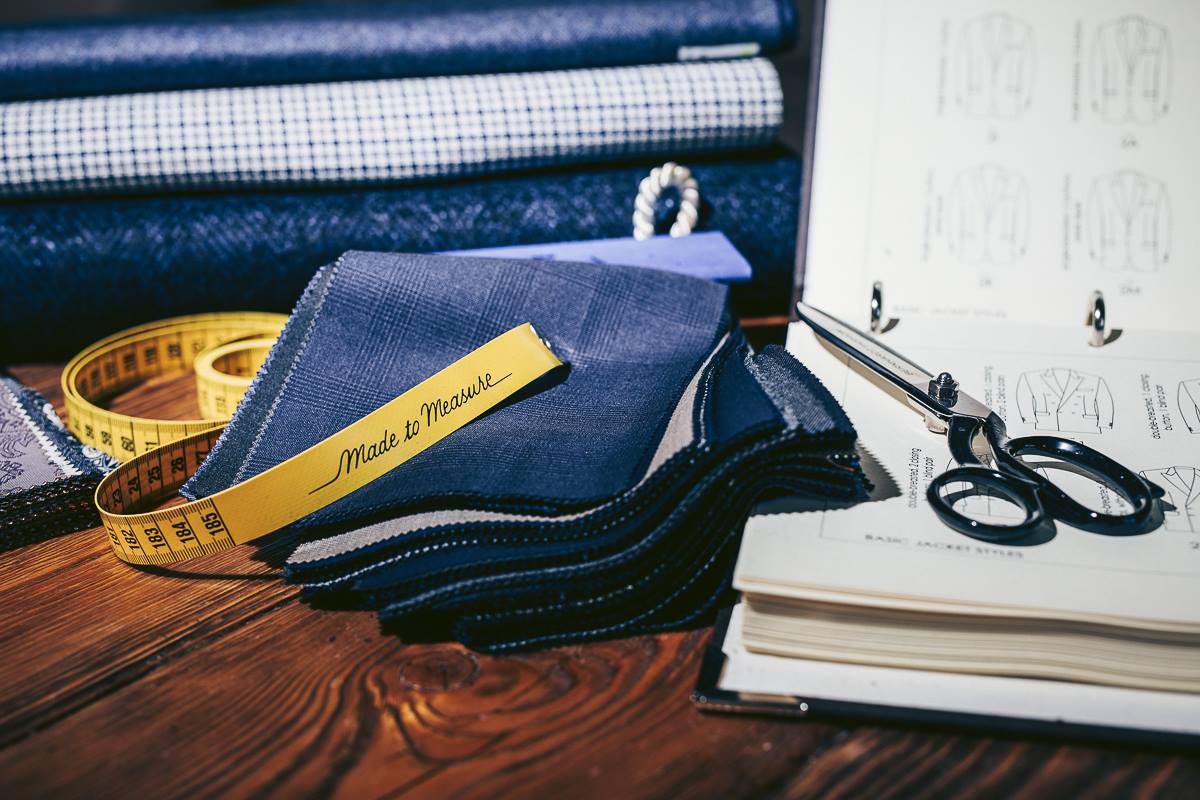 Cahier d'échantillons de tissus pour la confection de costumes sur mesure. On y voit des tissus de différentes teintes de bleu, un mètre de tailleur et des ciseaux de couture, sur une table de travail.