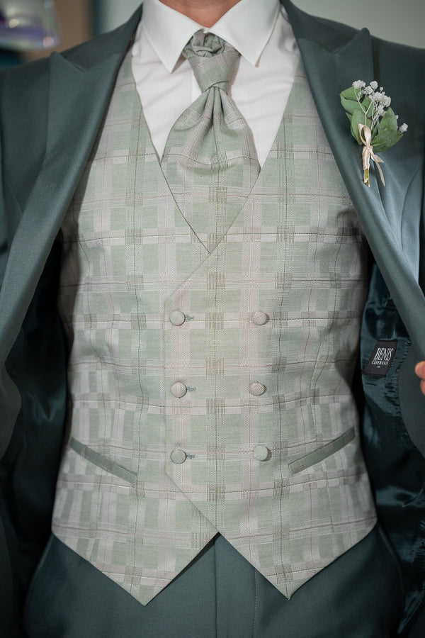 

L'image montre un homme portant un costume élégant. La veste est vert foncé et le gilet est vert clair à carreaux. La cravate est assortie au gilet. Le costume est de haute qualité et bien taillé. La boutonnière est ornée d'une petite fleur blanche. L'ensemble est très chic et parfait pour un mariage