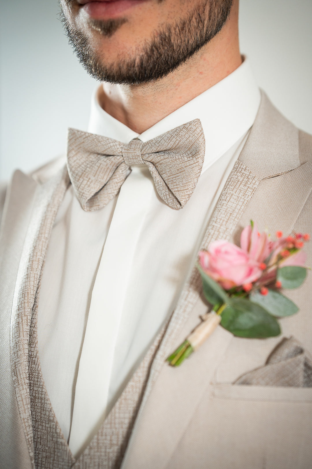 Détail du nœud papillon beige et la boutonnière fleur rose, du revers de col assorti au gilet avec un micro motifs géométrique ton sur ton beige