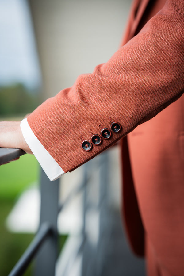 L'image montre un détail d'une veste de costume terracotta.  On voit principalement la manche, avec ses boutons foncés, et un bout de la chemise blanche portée en dessous. Le tissu de la veste semble avoir une texture légèrement texturée.  La photo est prise d'un angle qui met l'accent sur la texture et les détails du tissu et des boutons. L'arrière-plan est flou, ce qui attire l'attention sur les éléments principaux de l'image.  