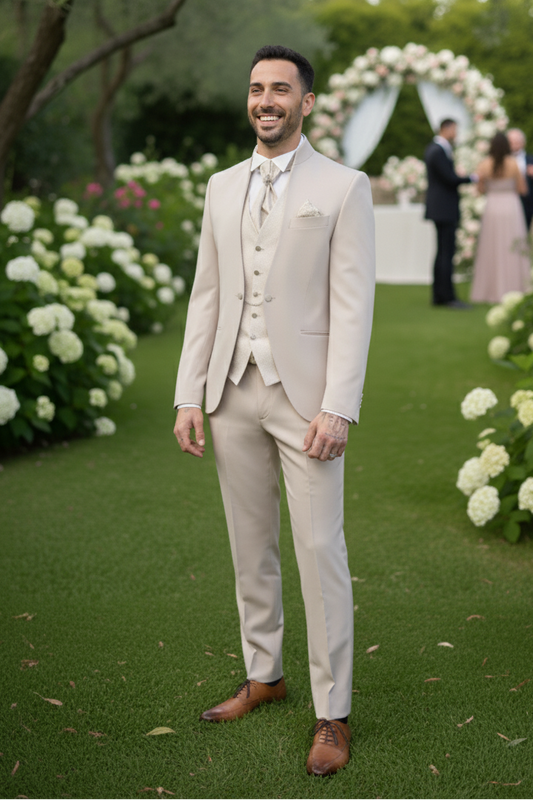 Photo d'un homme élégant, debout en plan pied, portant un costume trois-pièces de couleur beige ou crème (comme dans la photo de référence). Il sourit légèrement, se tient dans un jardin verdoyant avec des massifs de fleurs blanches (hortensias ou similaires) sur les côtés d'une allée de gazon, dans une ambiance de mariage extérieur. La lumière naturelle est douce, et l'arrière-plan flou suggère une arche de mariage floral
