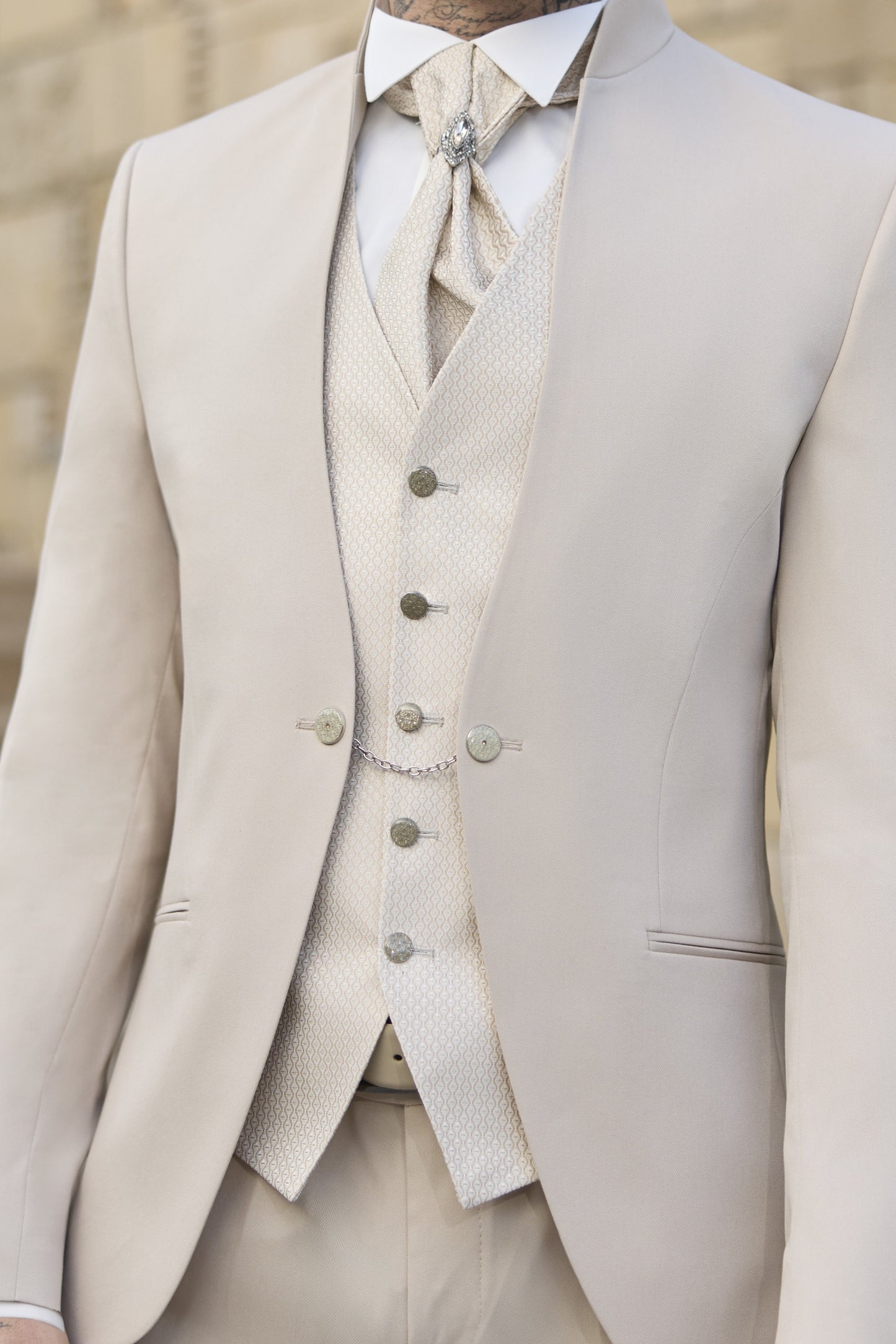 Gros plan sur un costume de mariage trois-pièces pour homme, de couleur beige clair ou crème, avec des détails élégants. Le gilet, de la même teinte, présente un motif texturé discret, et est orné d'une chaînette de montre gousset visible. La cravate ascot ou lavallière assortie est nouée avec une épingle décorative sertie de pierres. La veste ajustée, à col montant sans revers, montre des finitions impeccables. Parfait pour un marié élégant ou une cérémonie raffinée. Photographie éditoriale.