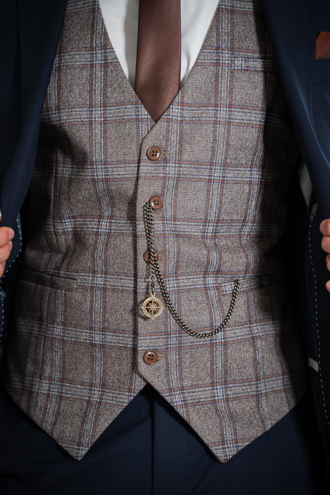 L'image montre un détail d'un gilet à carreaux brun et gris porté sur une chemise blanche et une cravate marron. Une montre de poche avec une chaîne est accrochée au gilet.  Le gilet est une pièce élégante et classique, qui évoque un stylerétro et vintage. La montre de poche ajoute une touche de sophistication et de tradition. L'ensemble suggère un homme attentif à son apparence et qui apprécie les détails.  